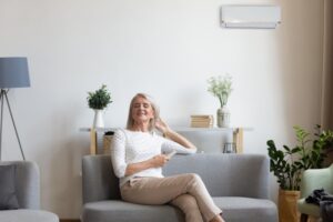 middle-aged-woman-sitting-on-sofa-smiling-with-mini-split-on-wall